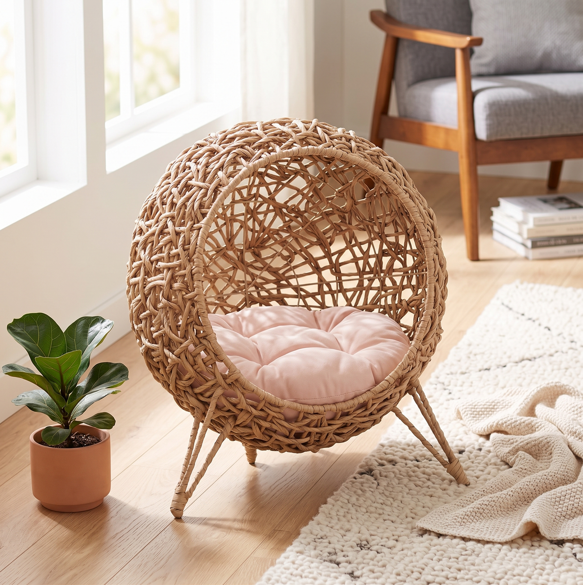 Handwoven water hyacinth egg-shaped cat bed with pink cushion, styled in a bright Scandinavian living room with wooden armchair and plant